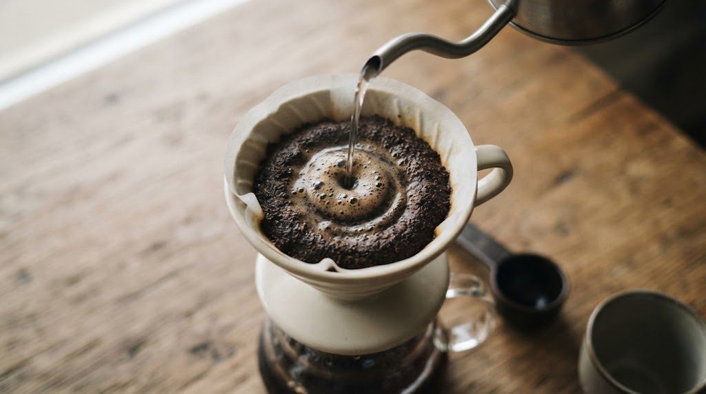 ケトルの細い注ぎ口から湯が注がれ、ドリッパーの中のコーヒー粉の中心から円を描くように泡が広がっている様子を斜め上から見たところ